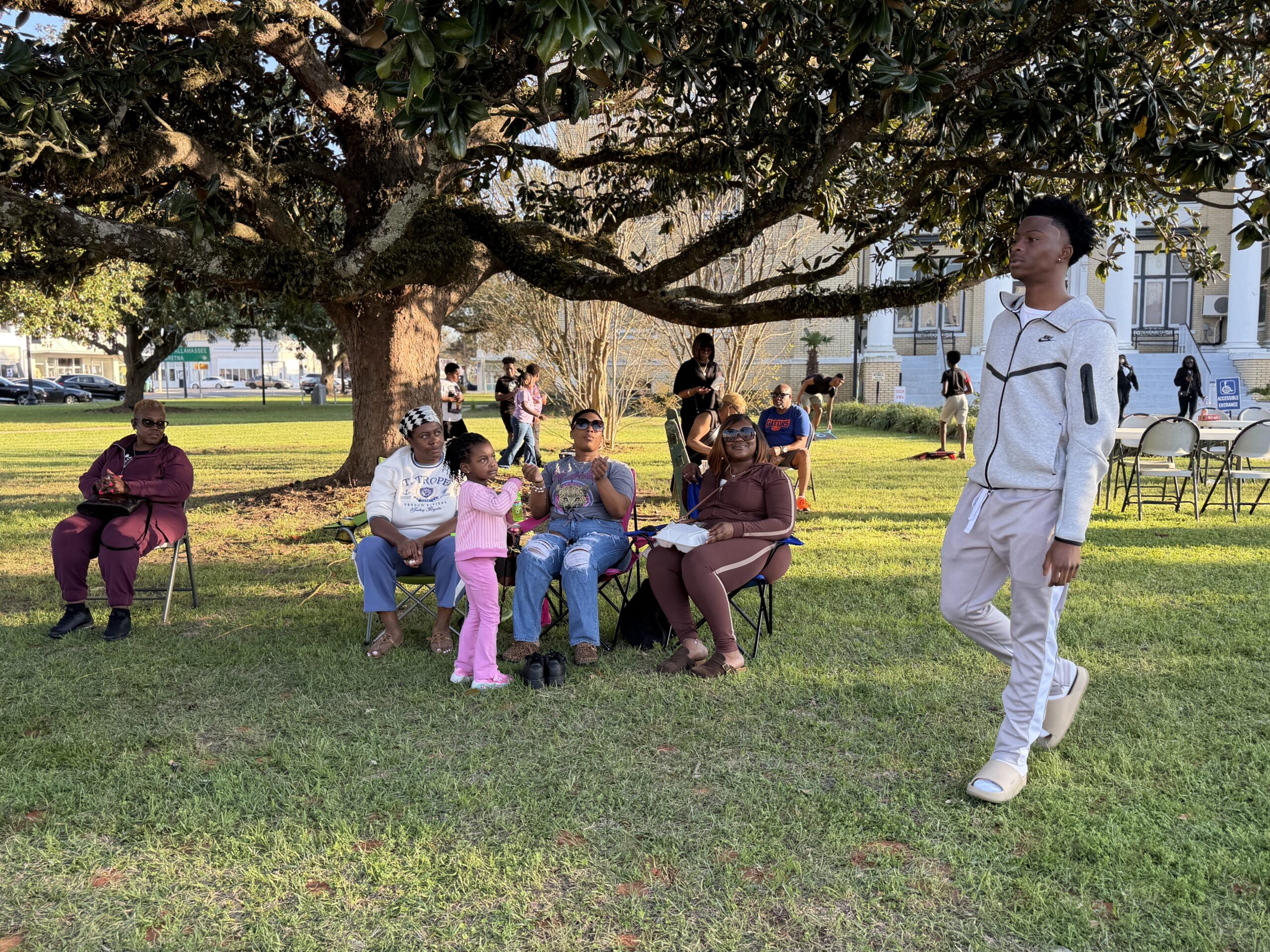 Families seated under a large tree at a park event while others walk and gather nearby during a community celebration.