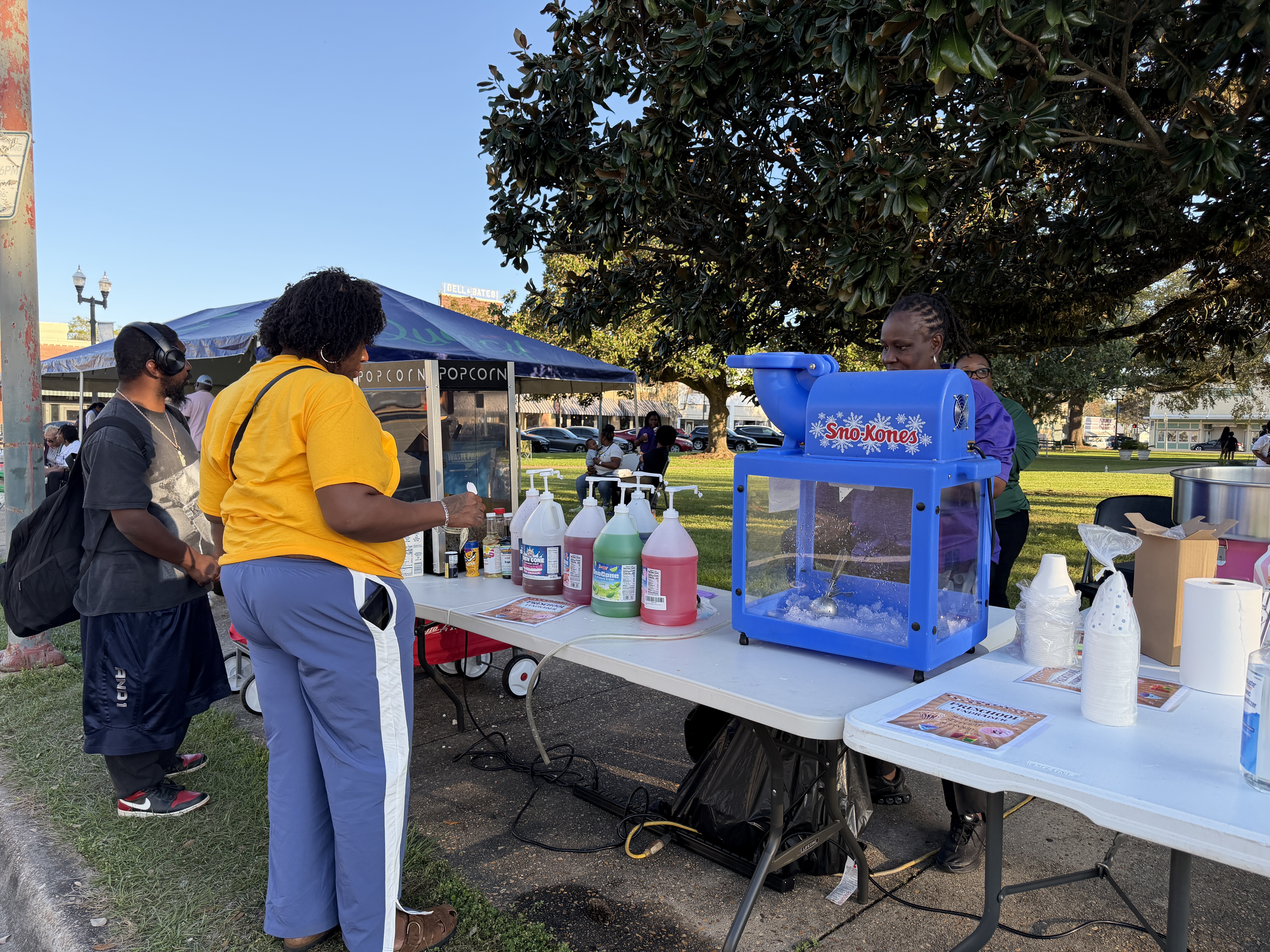 Community members gather at an outdoor event with a SnoKones stand serving flavored shaved ice under a large tree in a park setting.