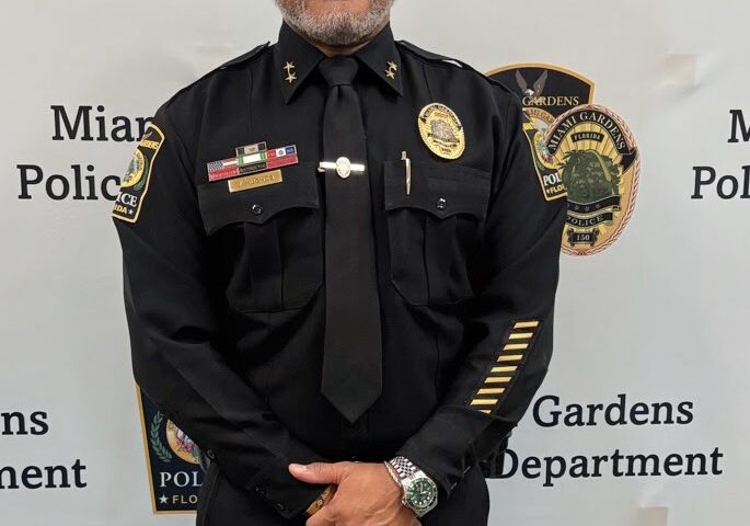 Portrait of a Miami Gardens Police Department officer in full uniform standing in front of a department backdrop displaying official insignia.