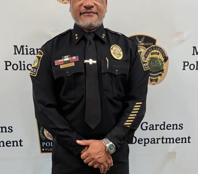 Portrait of a Miami Gardens Police Department officer in full uniform standing in front of a department backdrop displaying official insignia.