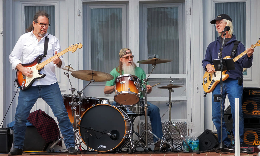 Three musicians performing live on an outdoor stage with electric guitar, drums, and bass during a Quincy Porchfest music performance.