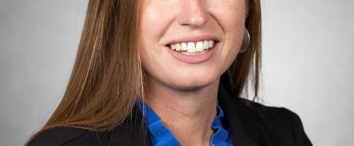 Professional headshot of a woman with long auburn hair wearing a black blazer and blue blouse, smiling against a neutral studio background.