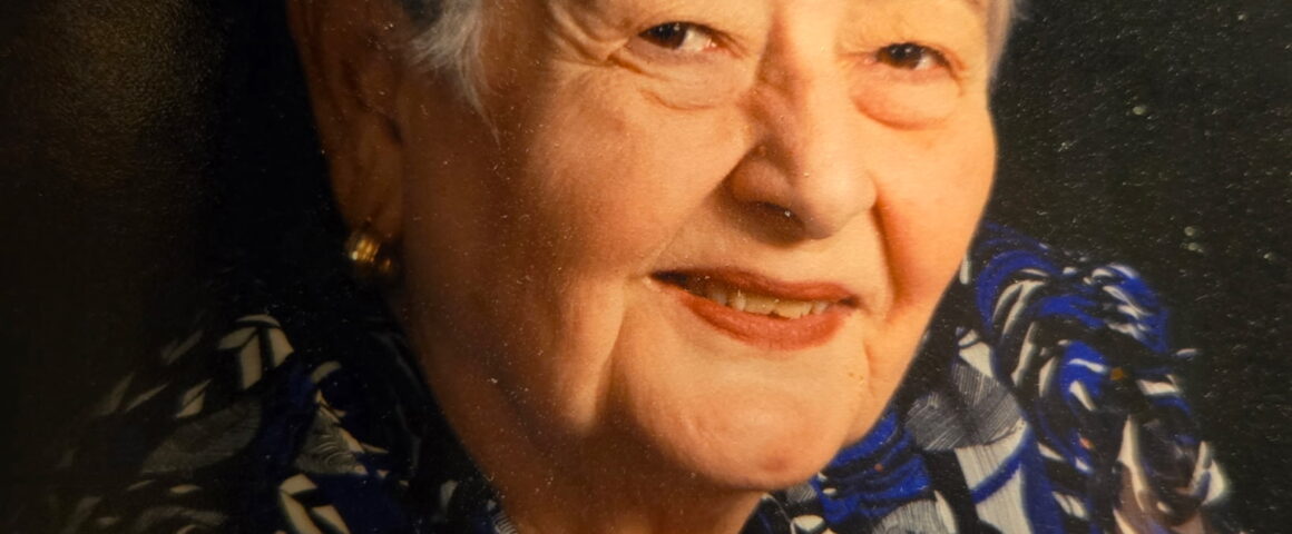 Portrait of an elderly woman with short gray hair wearing a patterned blouse, smiling against a dark studio background.