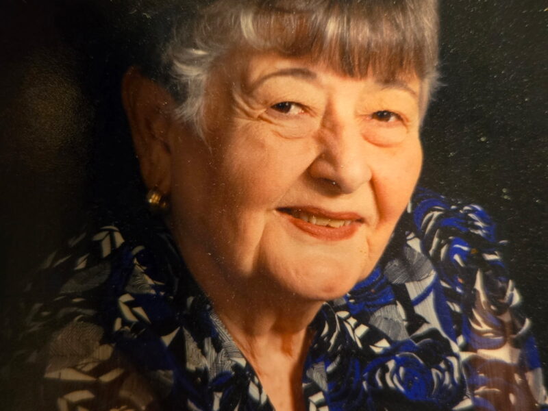 Portrait of an elderly woman with short gray hair wearing a patterned blouse, smiling against a dark studio background.