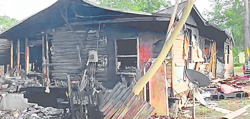 Severely burned house with heavy fire damage, charred siding, and debris scattered around the structure.