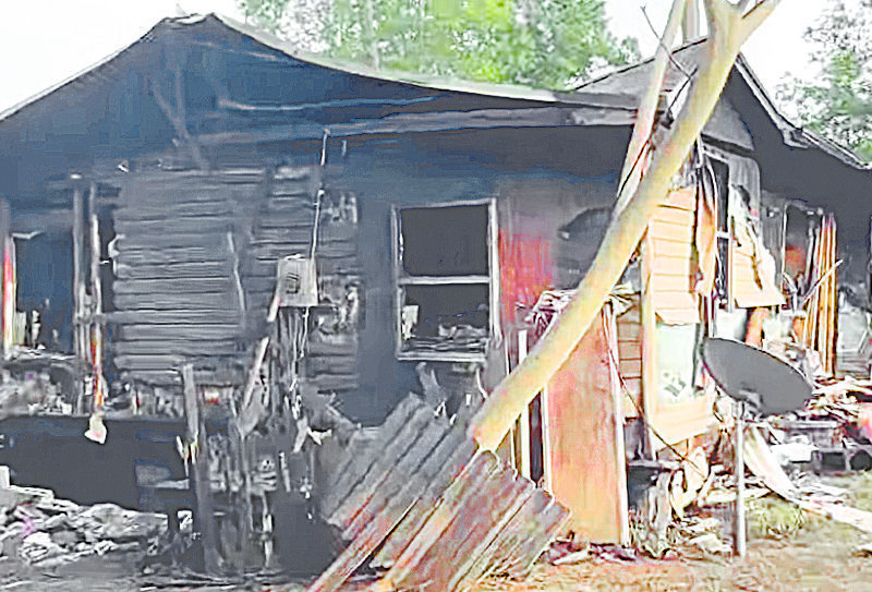 Severely burned house with heavy fire damage, charred siding, and debris scattered around the structure.
