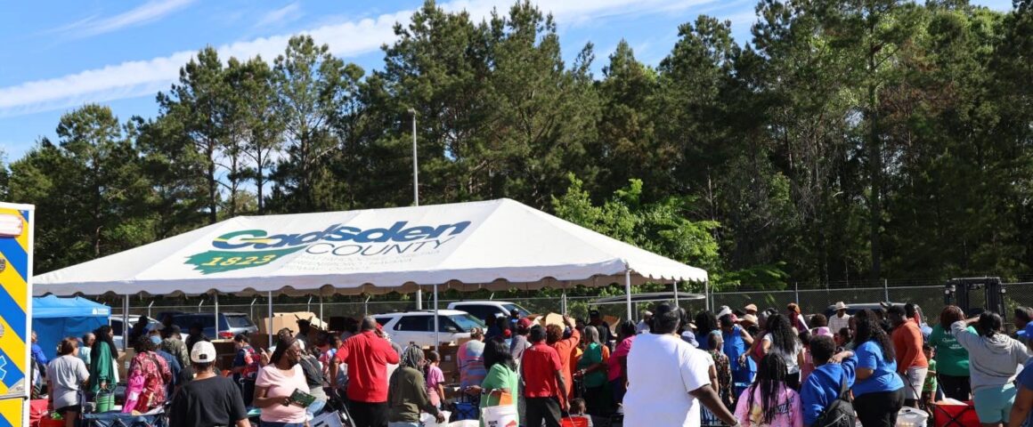 Large crowd gathered around a tent labeled Gadsden County, with people collecting items and pushing carts during a community distribution event.