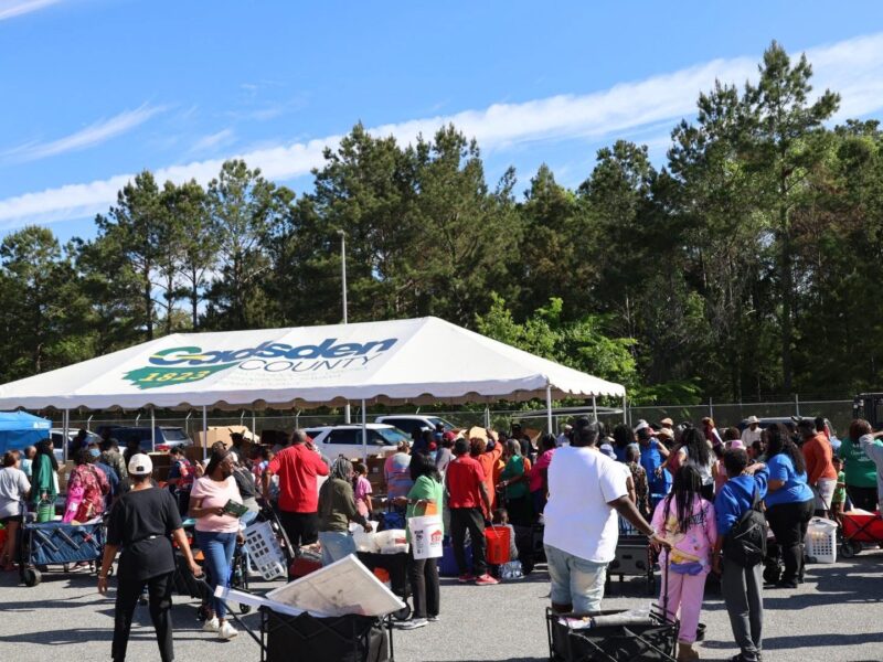 Large crowd gathered around a tent labeled Gadsden County, with people collecting items and pushing carts during a community distribution event.