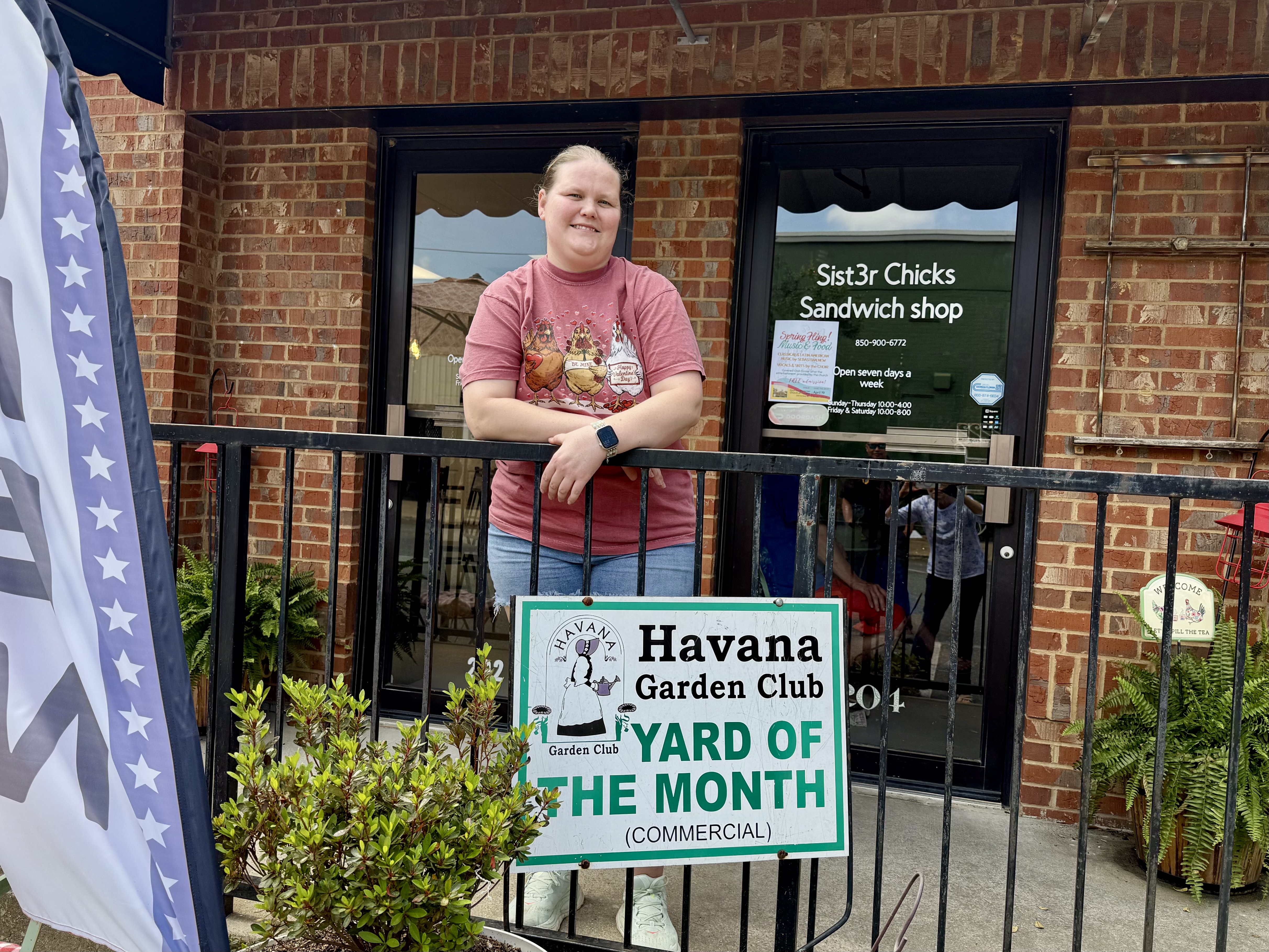 Kayla Edenfield standing outside Sist3r Chicks Sandwich Shop behind a sign reading “Havana Garden Club Yard of the Month (Commercial),” recognized as the April Commercial Yard of the Month.