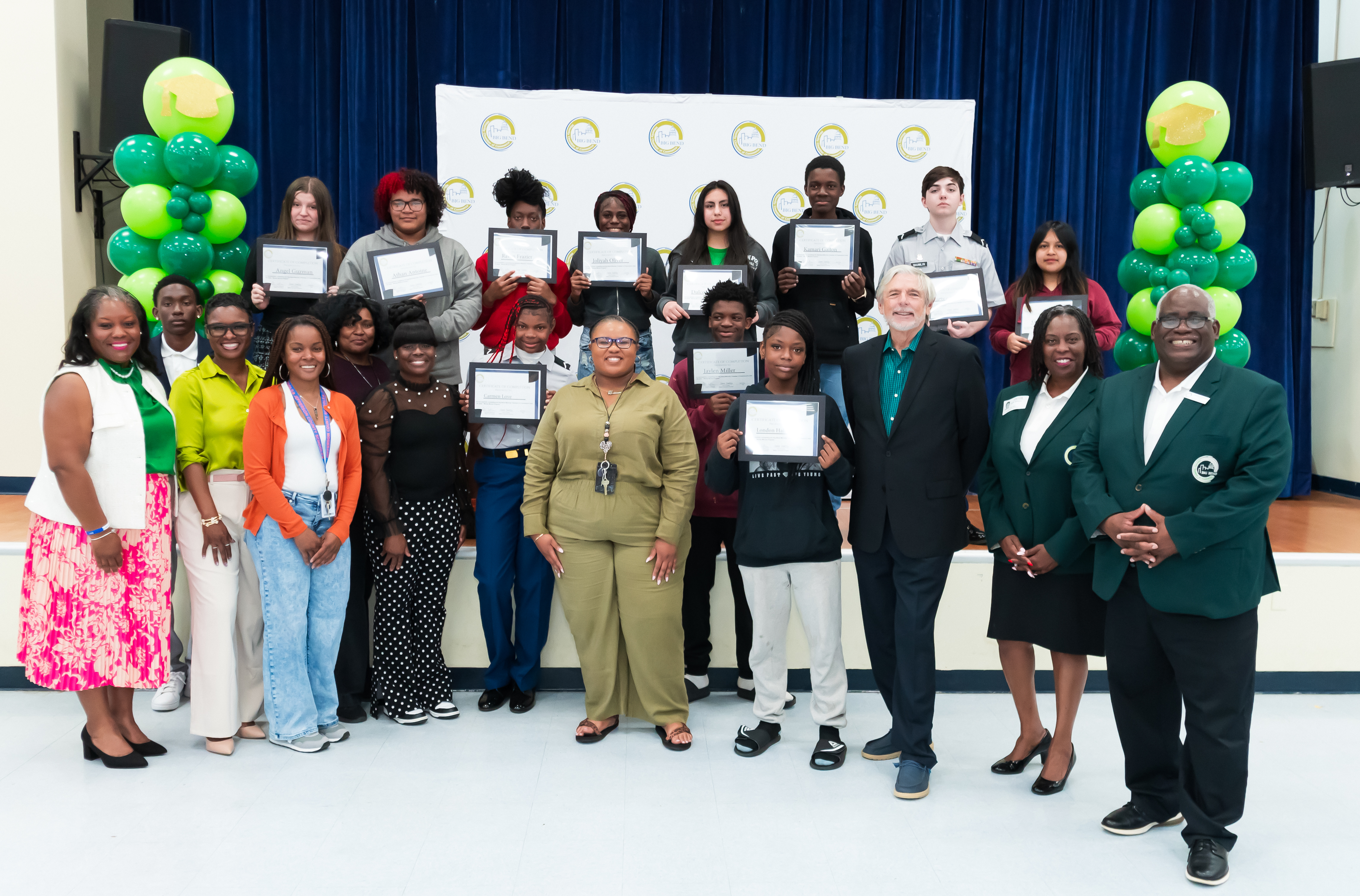 Group of students standing on a stage holding certificates, posing with adults and organizers in front of a backdrop and green balloon decorations at a graduation or recognition ceremony.