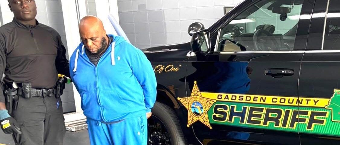 Man in a blue tracksuit standing in handcuffs next to a Gadsden County Sheriff’s patrol vehicle while a deputy stands nearby.