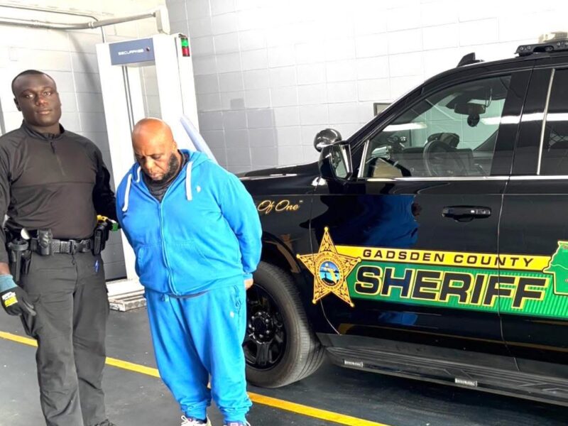 Man in a blue tracksuit standing in handcuffs next to a Gadsden County Sheriff’s patrol vehicle while a deputy stands nearby.