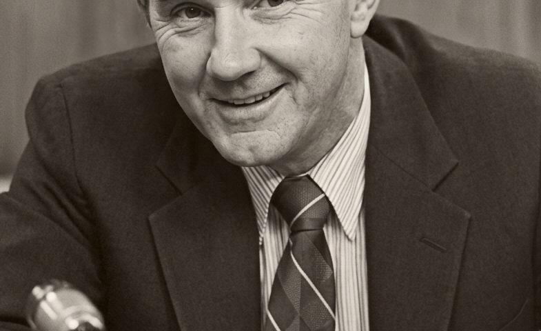 Black-and-white portrait of a man in a suit and patterned tie, smiling while seated at a desk with a microphone in the foreground.