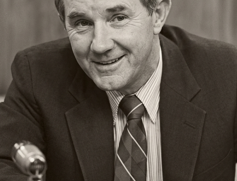 Black-and-white portrait of a man in a suit and patterned tie, smiling while seated at a desk with a microphone in the foreground.