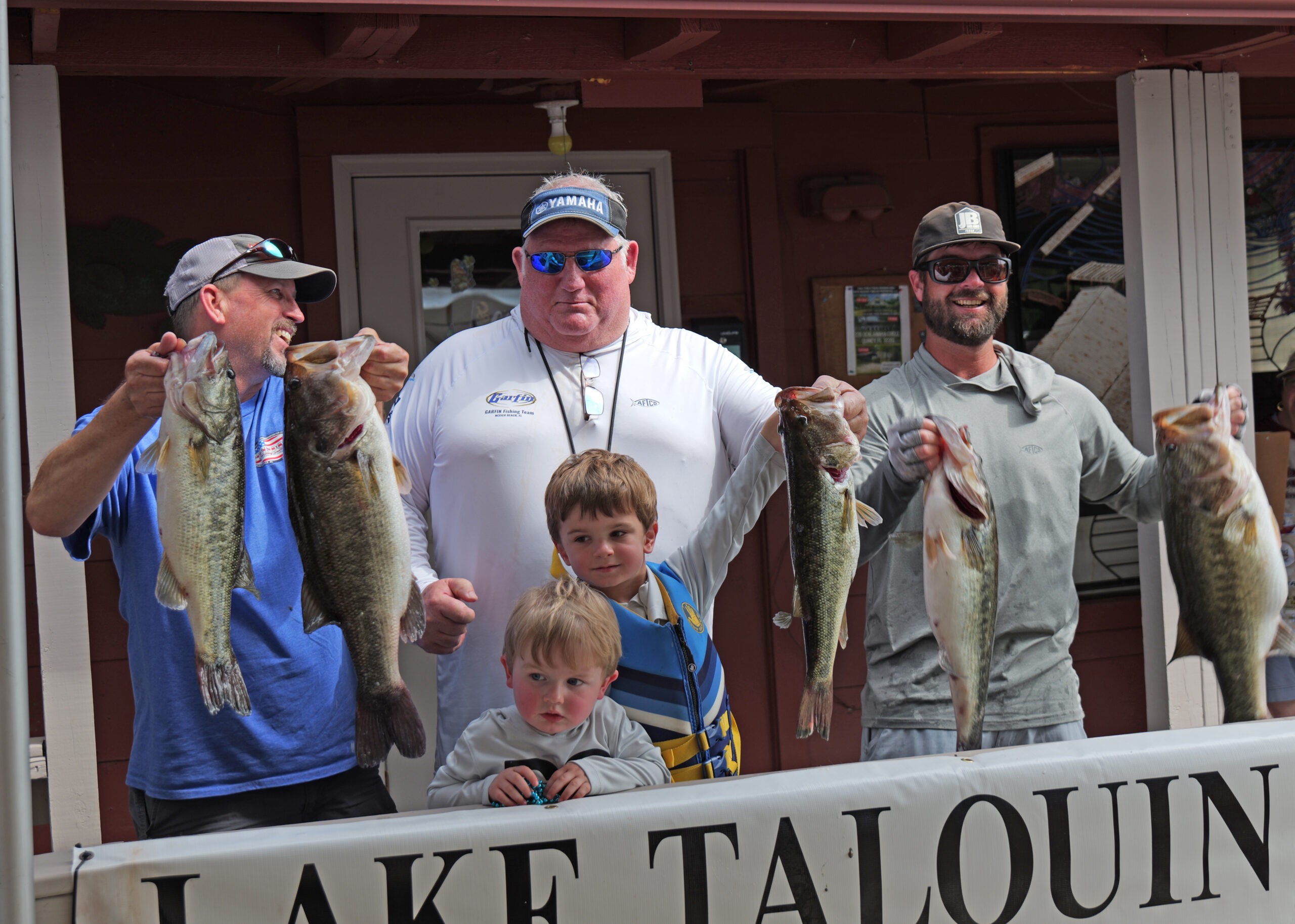 Four men and two young children pose in front of a “Lake Talquin” sign, holding several large bass fish, smiling after a successful fishing outing.