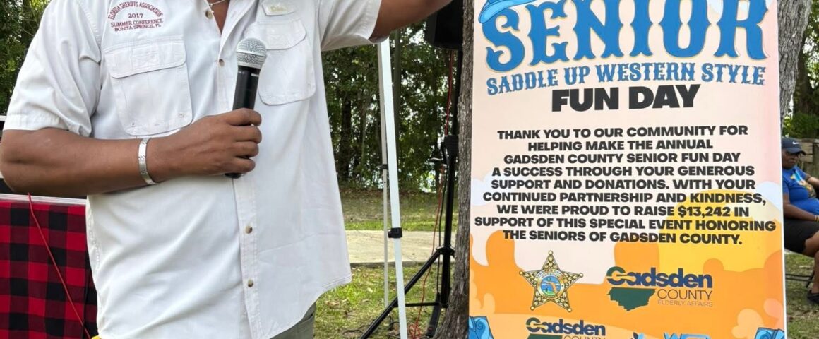 A man holding a microphone raises his hand beside a sign for the Senior Saddle Up Western Style Fun Day event outdoors beneath large trees.