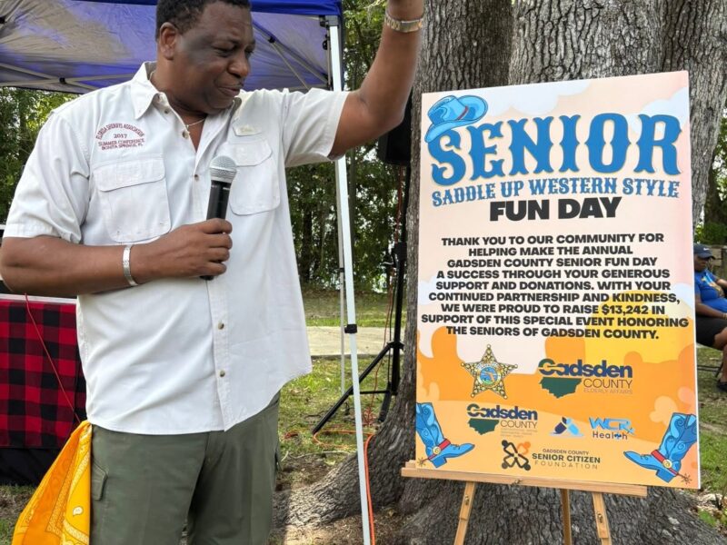 A man holding a microphone raises his hand beside a sign for the Senior Saddle Up Western Style Fun Day event outdoors beneath large trees.