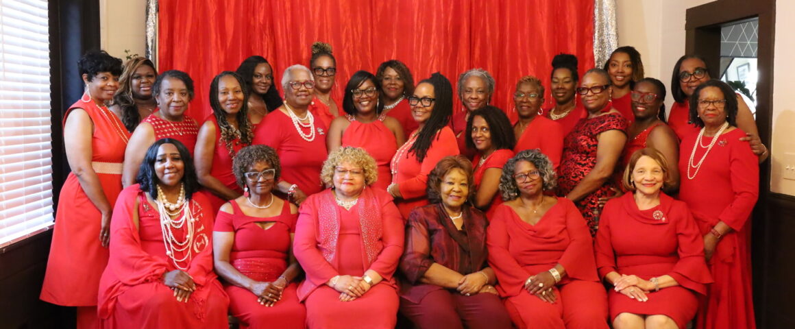 A large group photograph of more than twenty women dressed entirely in shades of red, burgundy, and pink posing together indoors. The group features a front row of seated women and two standing rows behind them, with many wearing pearl necklaces and jewelry. They are positioned in front of a bright red fabric backdrop framed by silver tinsel curtains.