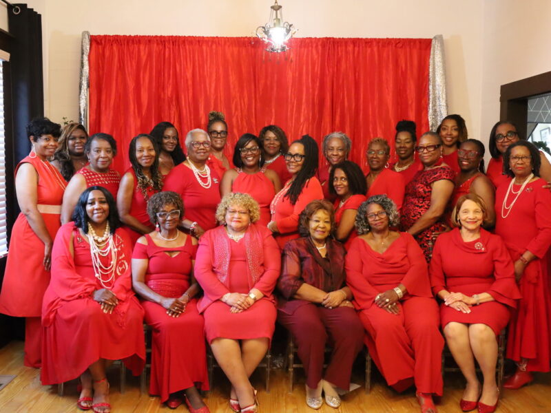 A large group photograph of more than twenty women dressed entirely in shades of red, burgundy, and pink posing together indoors. The group features a front row of seated women and two standing rows behind them, with many wearing pearl necklaces and jewelry. They are positioned in front of a bright red fabric backdrop framed by silver tinsel curtains.