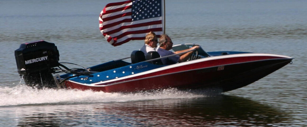 A red, white, and blue patriotic-themed speed boat cruising fast on the water for the Chattahoochee Thunder Nationals on the Apalachicola River. Two men are inside the boat, which features white stars on its blue trim, a large black Mercury outboard motor on the back, and a large American flag waving from a flagpole behind the seats.