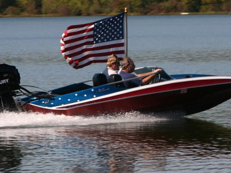 A red, white, and blue patriotic-themed speed boat cruising fast on the water for the Chattahoochee Thunder Nationals on the Apalachicola River. Two men are inside the boat, which features white stars on its blue trim, a large black Mercury outboard motor on the back, and a large American flag waving from a flagpole behind the seats.