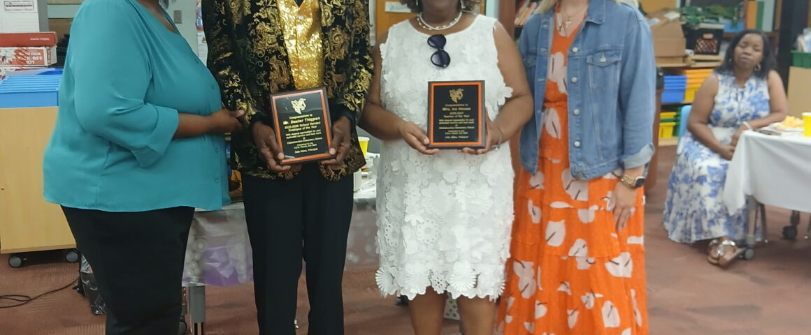 An awards presentation inside a school classroom, featuring four adults posing together in the foreground. A man dressed in a black hat, a black-and-gold patterned blazer, a gold sequined vest, and gold shoes stands next to a woman wearing a white lace dress and a white sun hat; both are holding black recognition plaques. They are flanked by two smiling women, one on the left in a teal blouse and black pants, and one on the right in an orange patterned dress and denim jacket.