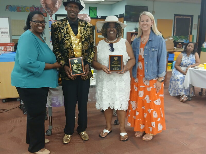 An awards presentation inside a school classroom, featuring four adults posing together in the foreground. A man dressed in a black hat, a black-and-gold patterned blazer, a gold sequined vest, and gold shoes stands next to a woman wearing a white lace dress and a white sun hat; both are holding black recognition plaques. They are flanked by two smiling women, one on the left in a teal blouse and black pants, and one on the right in an orange patterned dress and denim jacket.
