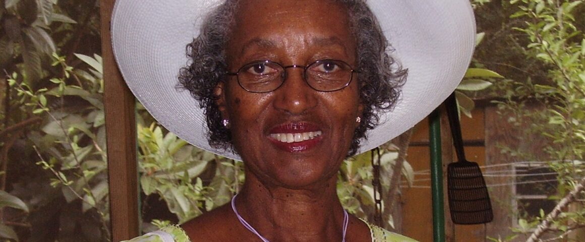 Obituary portrait of Elma Cummings. The photograph shows an older Black woman smiling warmly at the camera, wearing an elegant cream-colored dress or jacket and a matching wide-brimmed church hat with lace detailing against a neutral studio background.