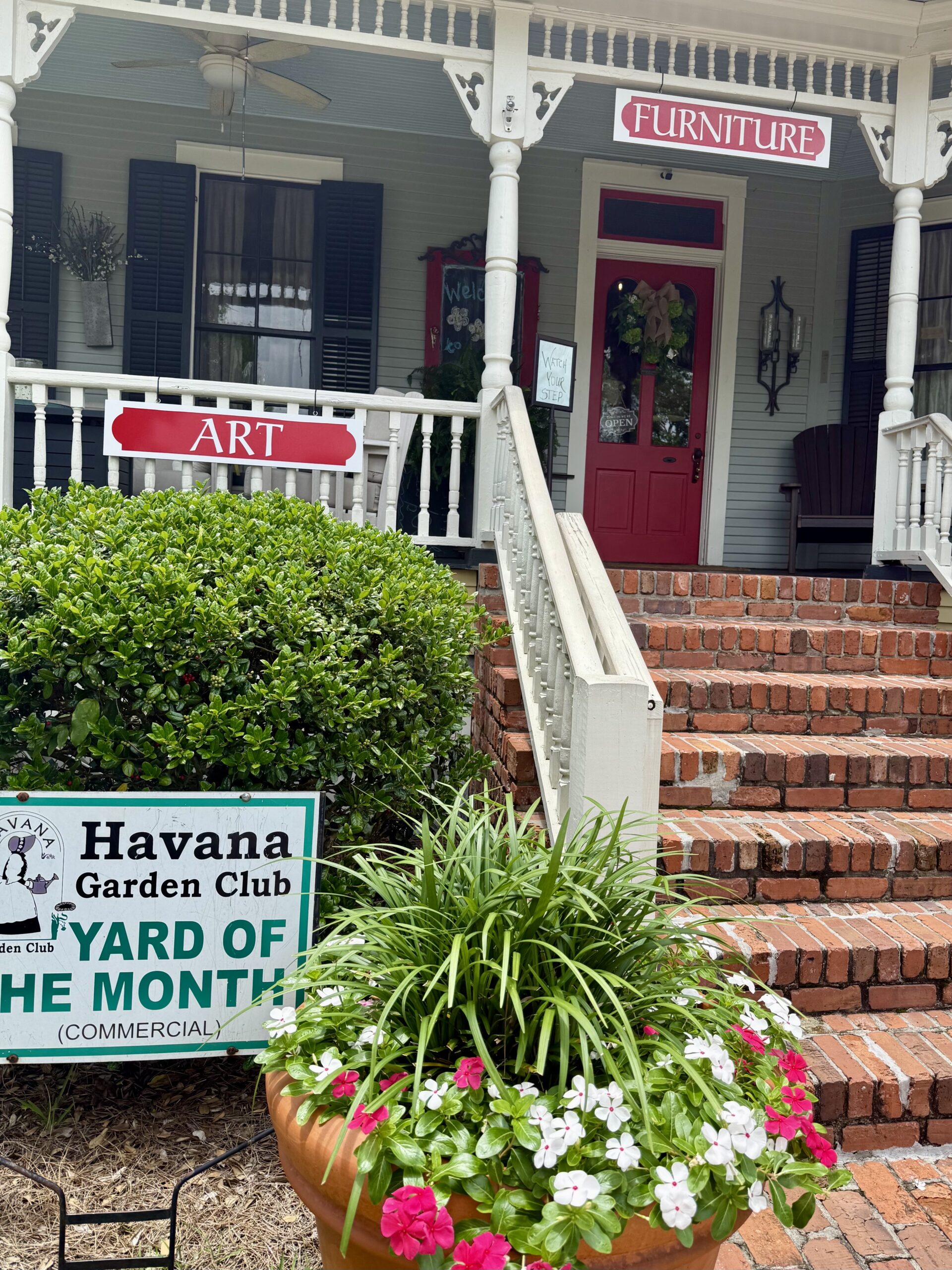 A brick stairway leads up to the covered porch of a commercial storefront business featuring a bright red door and decorative signs reading 'ART' and 'FURNITURE.' In the foreground, a planter filled with pink and white flowers sits next to a green lawn sign that reads 'Havana Garden Club Yard of the Month (Commercial).'