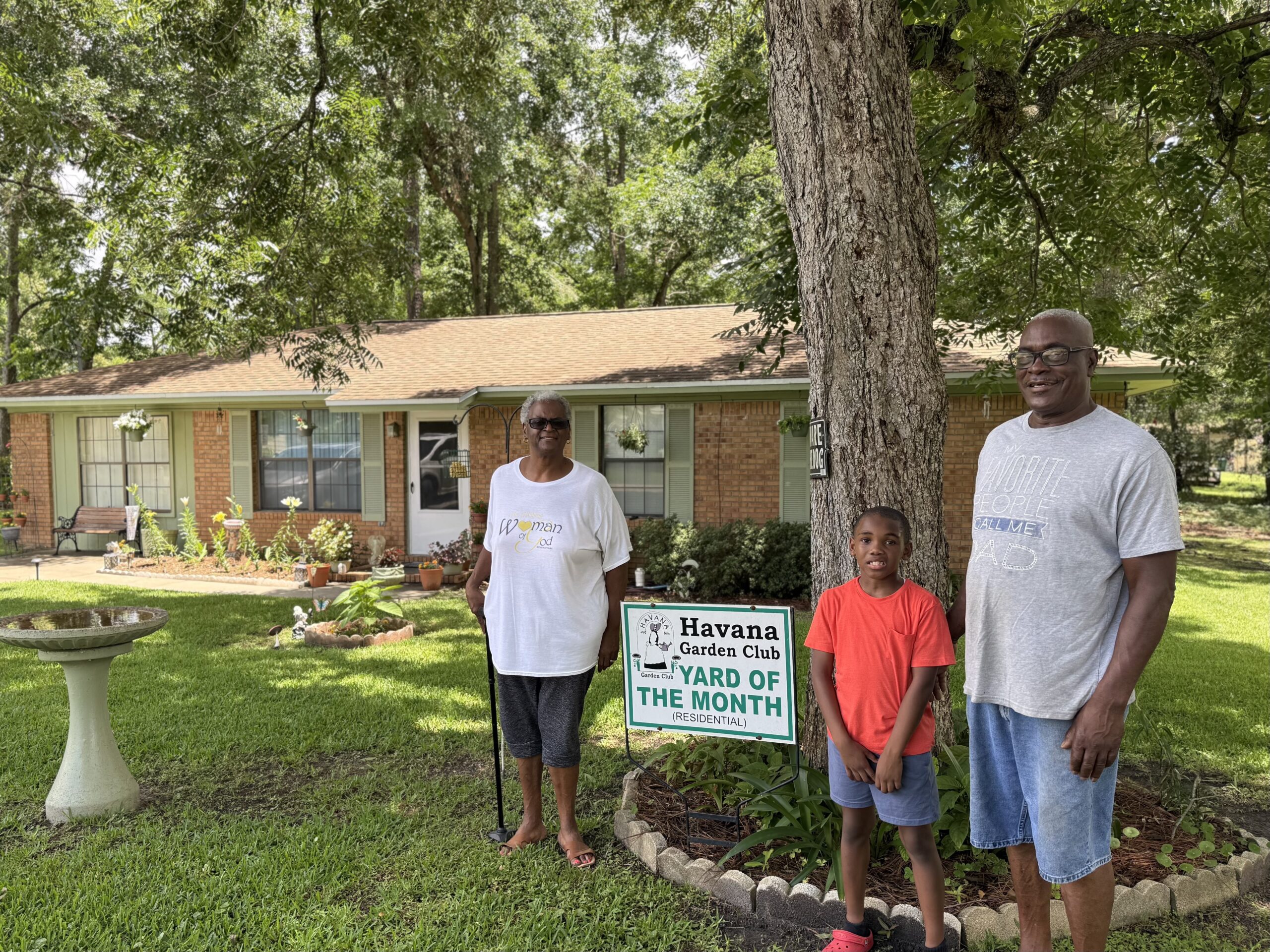 A woman, a young boy, and a man pose together on a green lawn in front of a sign that reads 'Havana Garden Club Yard of the Month (Residential).' The group stands next to a large tree on the right, with a well-landscaped, single-story brick ranch-style home, flower beds, and a concrete birdbath visible in the background.