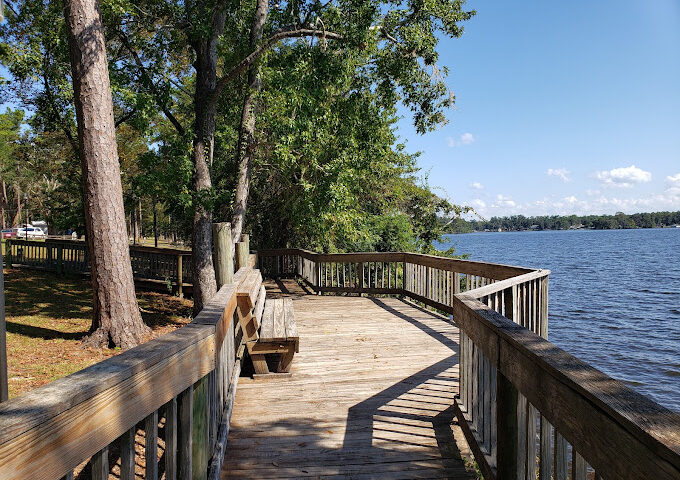A wooden boardwalk with handrails overlooks a wide, calm lake on a clear, sunny day. A wooden park bench is positioned along the walkway under the shade of mature trees lining the grassy shoreline. Shadows stretch across the wooden planks, and small white clouds dot the blue sky over the distant opposite shore.