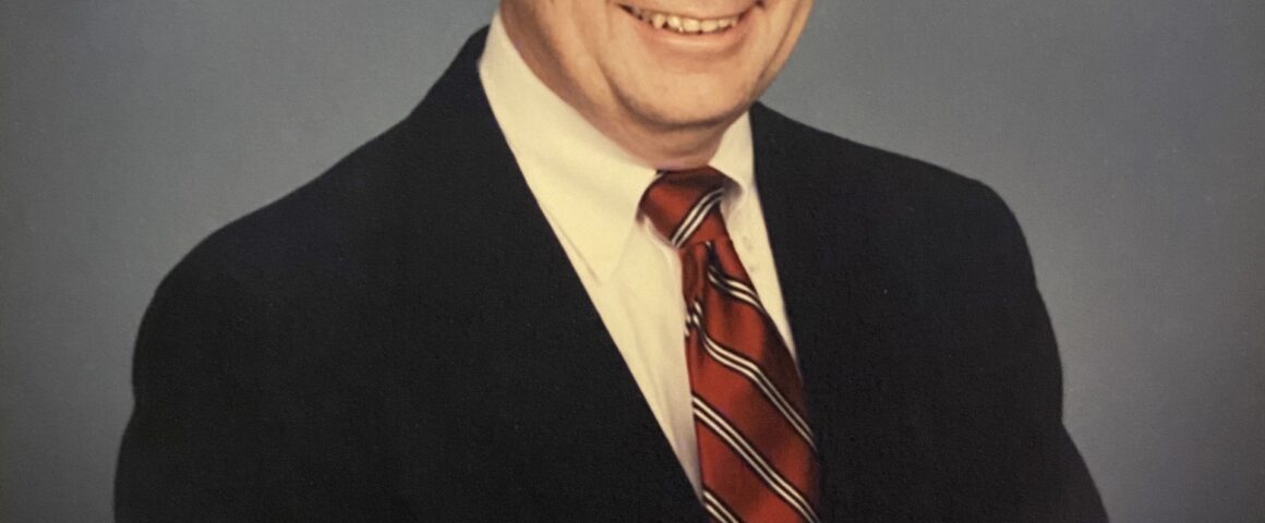 A professional studio headshot portrait of an older man smiling warmly. He has short, styled graying hair and is wearing a dark suit jacket, a white collared dress shirt, and a red tie with dark diagonal stripes. The photograph is shot from the chest up against a mottled blue-gray studio background, with an 'Olan Mills © 89' signature visible in the bottom-left corner.
