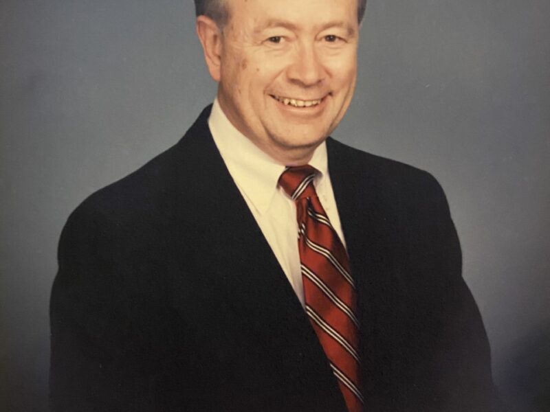 A professional studio headshot portrait of an older man smiling warmly. He has short, styled graying hair and is wearing a dark suit jacket, a white collared dress shirt, and a red tie with dark diagonal stripes. The photograph is shot from the chest up against a mottled blue-gray studio background, with an 'Olan Mills © 89' signature visible in the bottom-left corner.