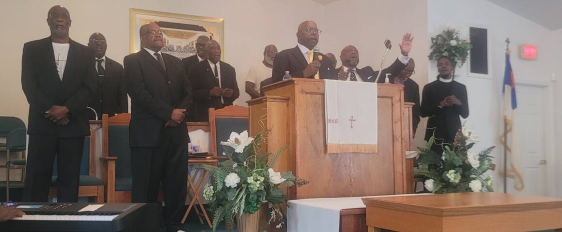 Reverend Danny Whitfield preaches from a central wooden pulpit with one hand raised at Bear Creek Bethel Missionary Baptist Church during an 'Iron Sharpens Iron' event. He is surrounded by several men in dark suits standing on the stage, including Pastor Terrence D. Milton to the left of the pulpit. The sanctuary stage features white floral arrangements, an altar table, a crystal chandelier hanging above, and musical equipment in the foreground.