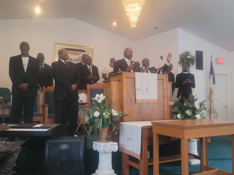 Reverend Danny Whitfield preaches from a central wooden pulpit with one hand raised at Bear Creek Bethel Missionary Baptist Church during an 'Iron Sharpens Iron' event. He is surrounded by several men in dark suits standing on the stage, including Pastor Terrence D. Milton to the left of the pulpit. The sanctuary stage features white floral arrangements, an altar table, a crystal chandelier hanging above, and musical equipment in the foreground.