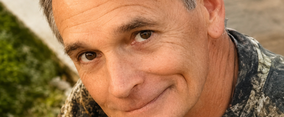 Obituary portrait of William Benjamin “Ben” Murray. The close-up photograph shows a middle-aged man with short graying hair smiling gently at the camera. He is wearing a camouflage print shirt against a blurred outdoor background.