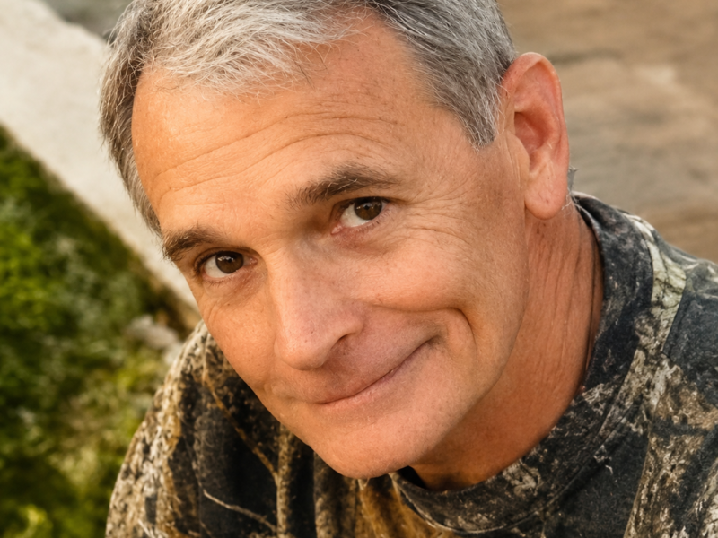 Obituary portrait of William Benjamin “Ben” Murray. The close-up photograph shows a middle-aged man with short graying hair smiling gently at the camera. He is wearing a camouflage print shirt against a blurred outdoor background.