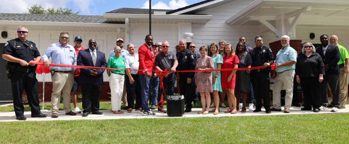 A large group of local officials, police officers, and community members gathered outside a white single-story building for a ribbon-cutting ceremony under a blue sky. Two police officers hold a long red ribbon across the front lawn while a man in a red suit jacket stands near the center next to others holding giant scissors.