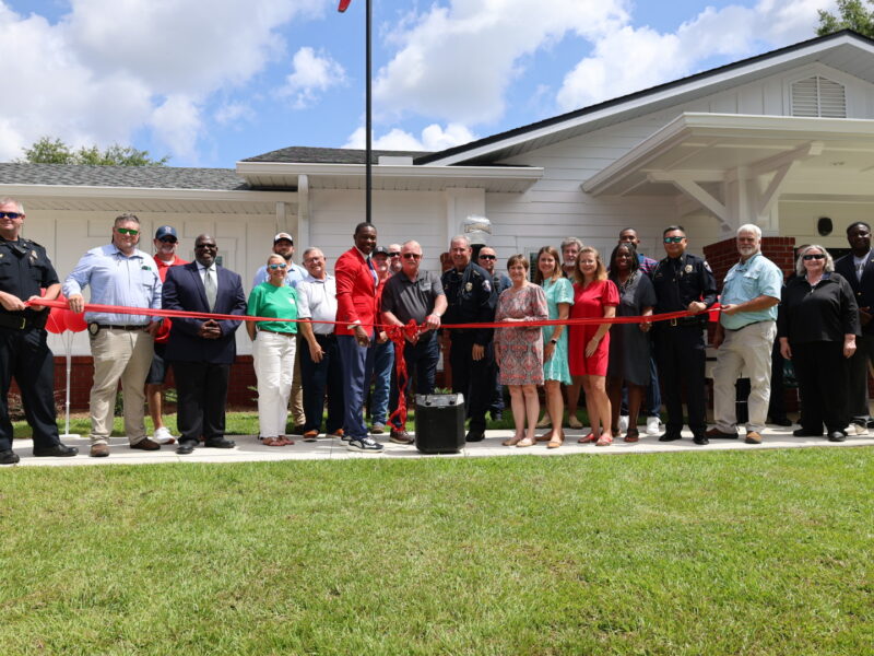 A large group of local officials, police officers, and community members gathered outside a white single-story building for a ribbon-cutting ceremony under a blue sky. Two police officers hold a long red ribbon across the front lawn while a man in a red suit jacket stands near the center next to others holding giant scissors.