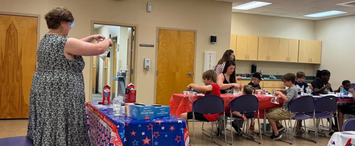 An indoor community room scene where several children and adults are seated at long tables making homemade ice cream. The tables are decorated with red, white, and blue patriotic tablecloths. On the left, an instructor in a patterned dress prepares supplies on a utility table holding milk cartons and a blue box of storage bags.