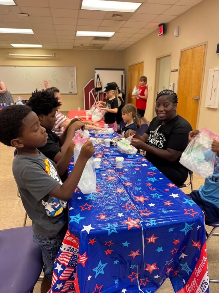 A group of children and an adult woman sitting along a table covered in a blue 'Happy America 250!' star-patterned tablecloth. They are actively handling clear plastic baggies containing ice and cream mixture to make baggie ice cream. A smiling Black woman in a black t-shirt looks directly toward the camera.
