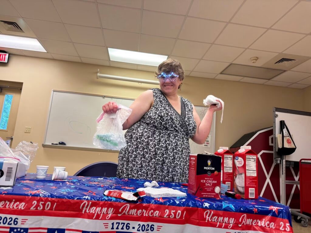 A woman in a black and white sleeveless patterned dress smiling playfully while holding up a clear plastic baggie containing an ice cream mixture. She is wearing blue, glowing light-up star-shaped glasses. In front of her on a patriotic 'Happy America 250!' tablecloth sit a red box of Morton Rock Salt and cartons of heavy whipping cream.