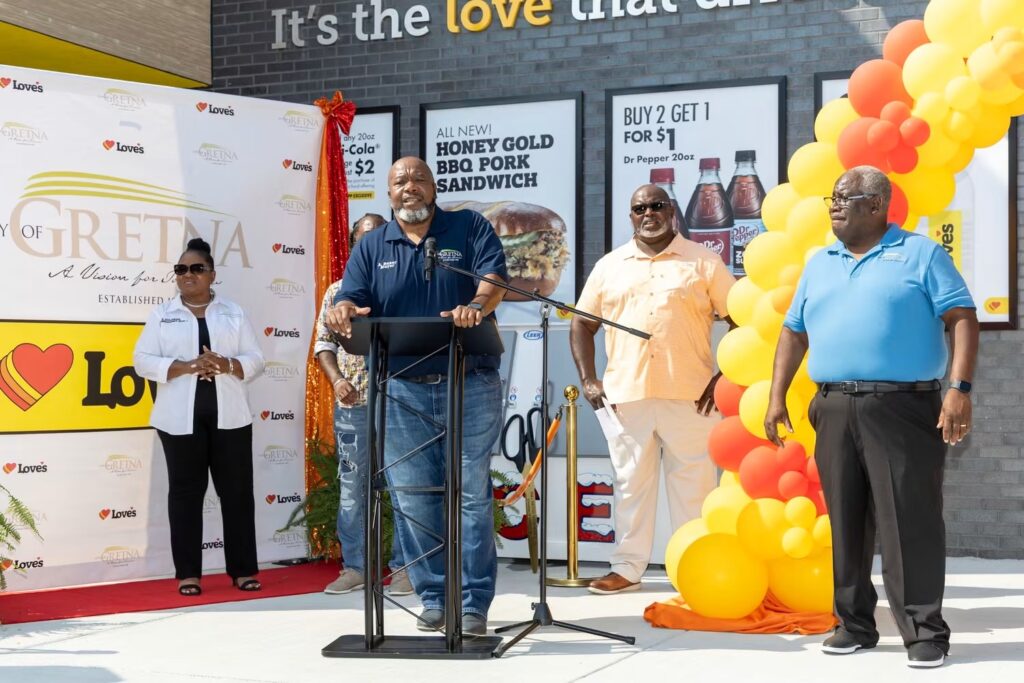 A man stands at a black podium speaking into a microphone during a grand opening event outside a Love's Travel Stop. He is wearing a navy blue polo shirt and jeans. Several other individuals stand nearby on a red carpet area, next to a large column of yellow and orange balloons and promotional posters for food items.