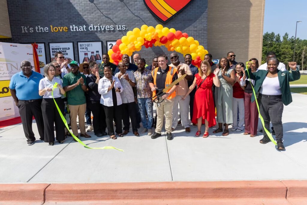 A large group of community members and officials gather for a ribbon-cutting ceremony outside a newly built Love's Travel Stop. In the center, a young man in a high-visibility orange vest holds giant ceremonial scissors, while attendees on both sides hold up a long, bright yellow ribbon, cheering and clapping under a yellow and orange balloon arch.