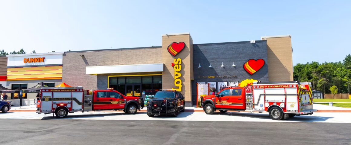 A wide exterior shot of a modern Love's Travel Stop and attached Dunkin' storefront on a clear day. Parked in a line across the asphalt parking lot out front are three emergency vehicles: a red Midway Fire Rescue truck, a black police utility vehicle, and a red City of Gretna Fire Rescue truck.