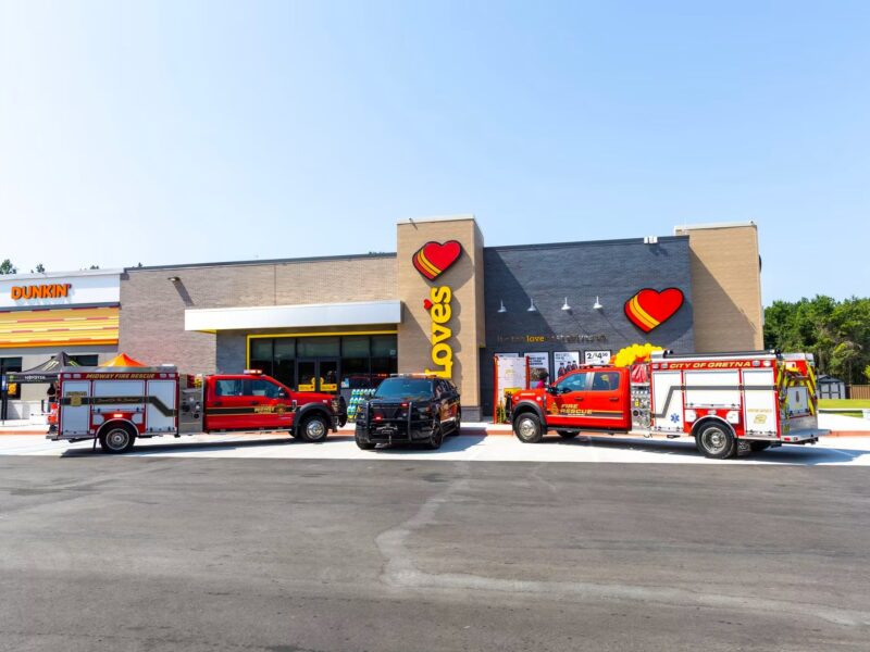 A wide exterior shot of a modern Love's Travel Stop and attached Dunkin' storefront on a clear day. Parked in a line across the asphalt parking lot out front are three emergency vehicles: a red Midway Fire Rescue truck, a black police utility vehicle, and a red City of Gretna Fire Rescue truck.