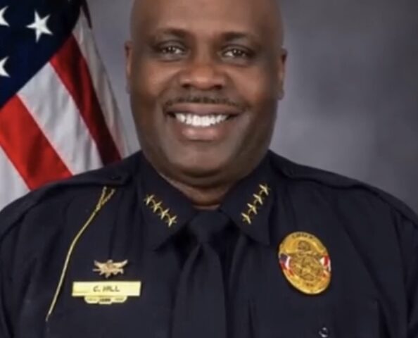 A professional studio headshot of a Black male police officer, Chief C. Hill, smiling broadly at the camera. He is dressed in a dark blue law enforcement uniform featuring four gold stars on each collar flap, a gold badge on his chest, and a gold name tag that reads 'C. HILL' against a gray background with an American flag on the left.