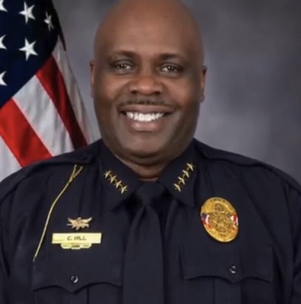A professional studio headshot of a Black male police officer, Chief C. Hill, smiling broadly at the camera. He is dressed in a dark blue law enforcement uniform featuring four gold stars on each collar flap, a gold badge on his chest, and a gold name tag that reads 'C. HILL' against a gray background with an American flag on the left.