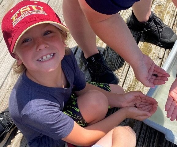 A close-up, high-angle shot of a young boy smiling broadly at the camera on a wooden dock. He is wearing a red Florida State University (FSU) baseball cap and a dark blue t-shirt, holding out his cupped hands containing small aquatic organisms alongside other hands.