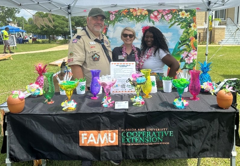Three people standing behind a display table under a white pop-up tent at a grassy outdoor fair. The table features a black tablecloth with the 'FAMU Cooperative Extension' logo and is decorated with bright neon tropical cocktail glasses and floral leis. A tropical beach-themed backdrop hangs behind them.